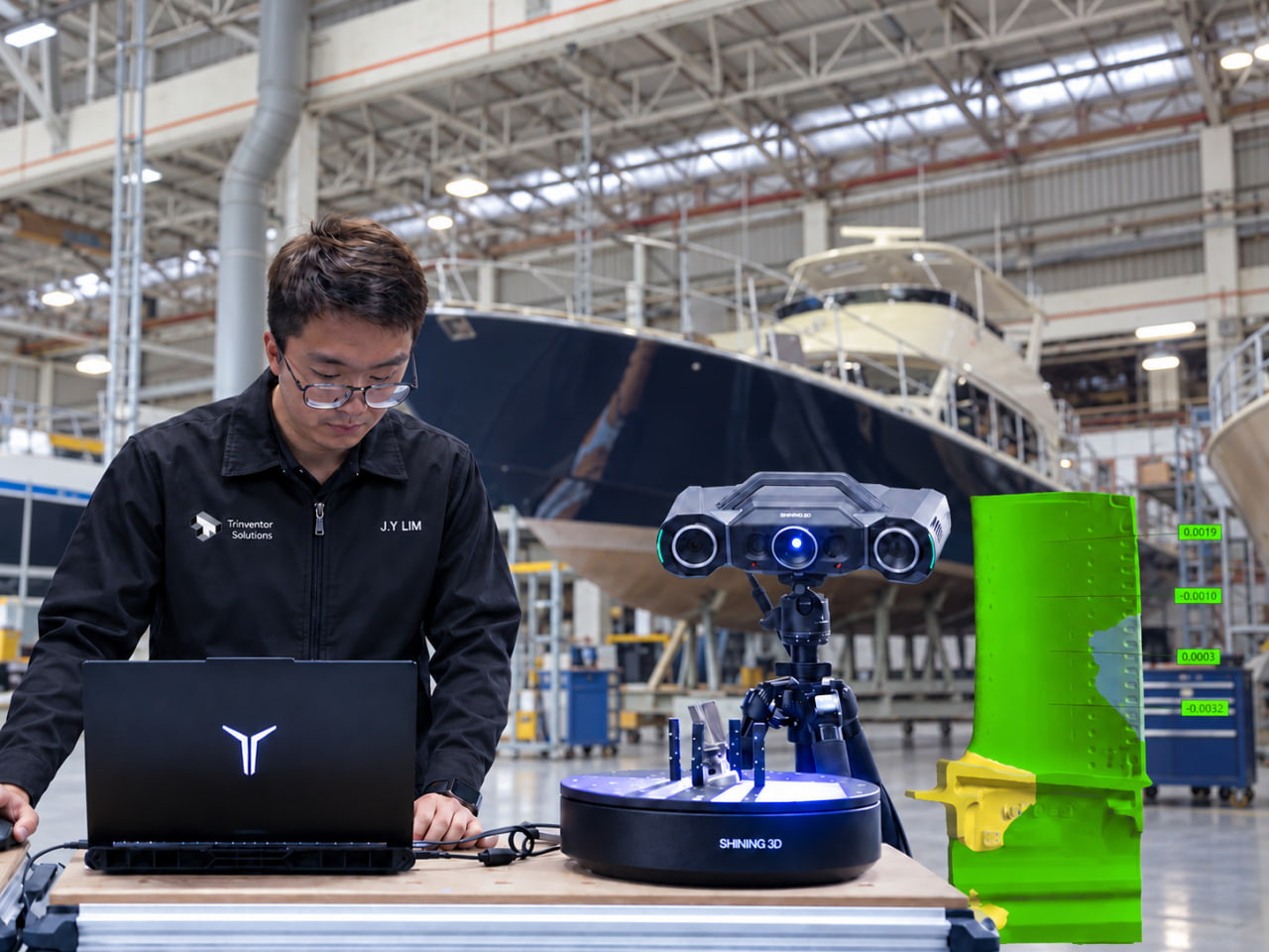 Engineer at a laptop beside a 3D scanner on a tripod and a turntable holding machined parts, with a deviation map on screen