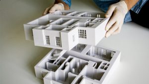 A detailed 3D printed architectural model being lifted in gloved hands, showing interior walls and window mullions