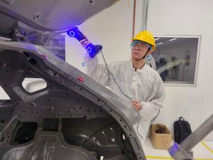 Handheld 3D scanner measuring a bare car body-in-white on a workshop line, with reflective markers on the frame rail