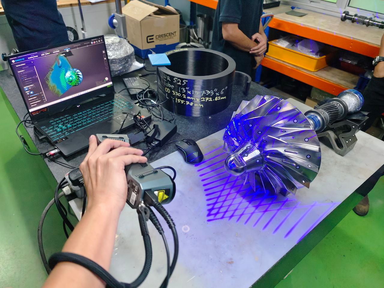 Handheld 3D scanner projecting blue laser lines across a machined impeller on a workbench, with the live mesh on a laptop