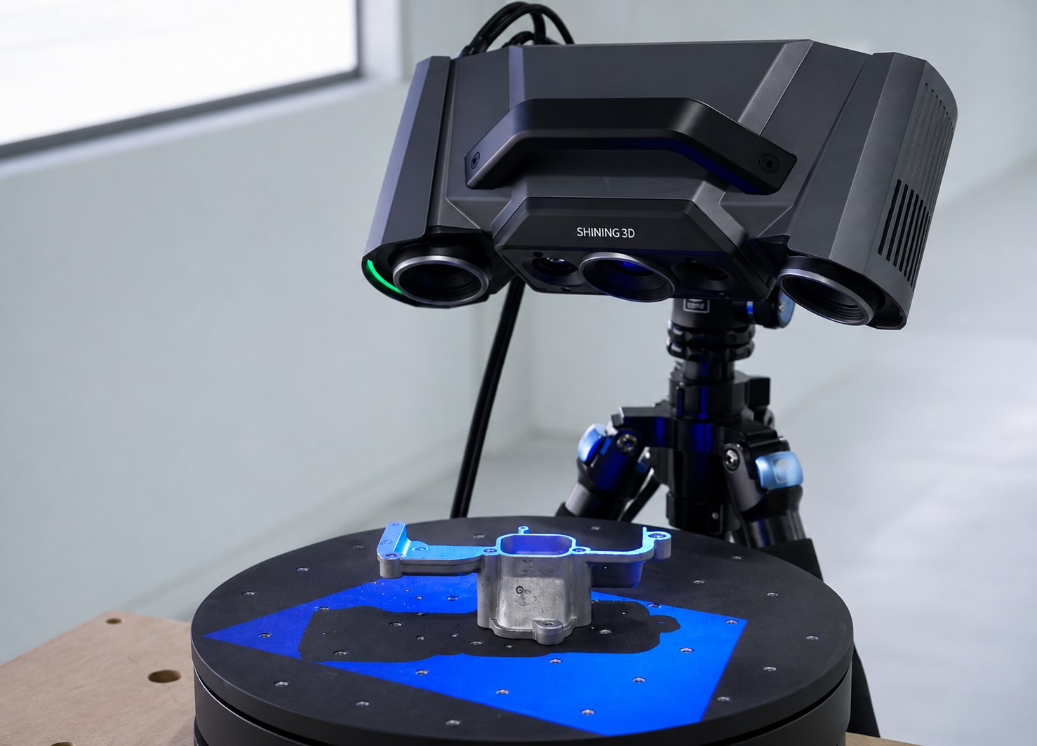 Structured-light 3D scanner on a tripod above a black rotary turntable holding a machined bracket in a laboratory scanning setup