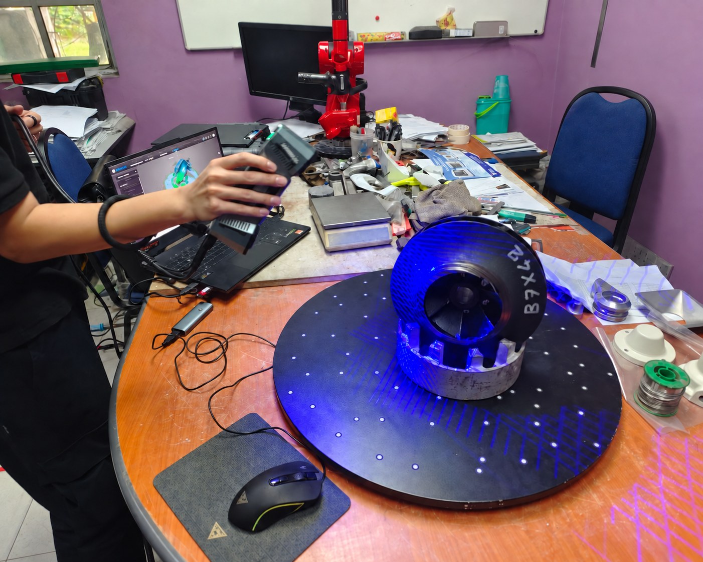 Rotary turntable with a machined part on it being scanned by a handheld 3D scanner beside a laptop