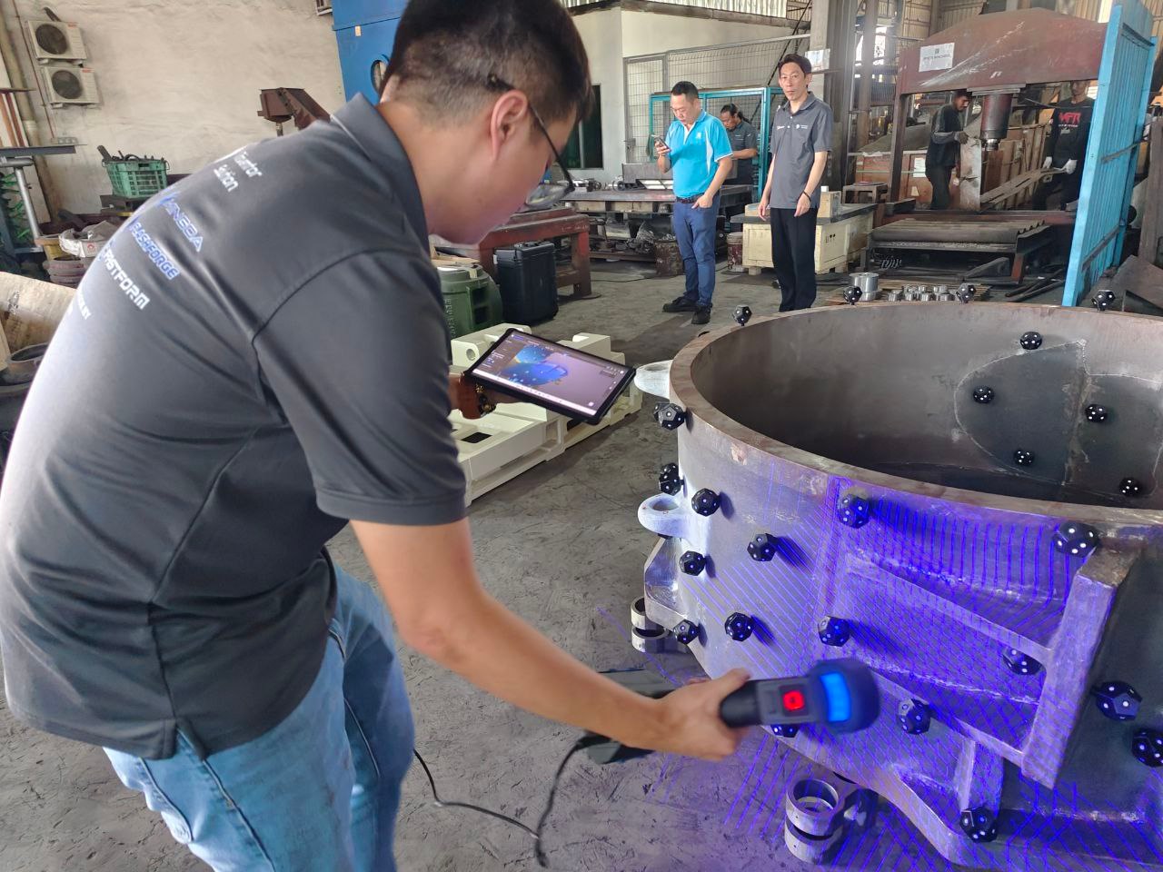 Handheld 3D scanner sweeping blue laser lines across a large fabricated steel ring on a workshop floor