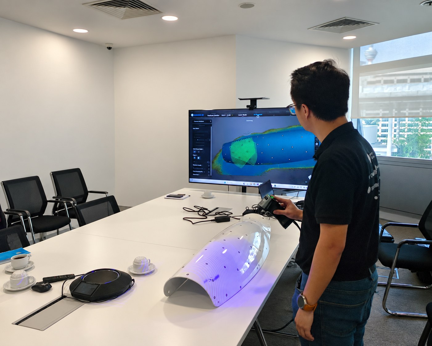 Handheld 3D scanner capturing a large curved panel on a conference table with the scan on screen