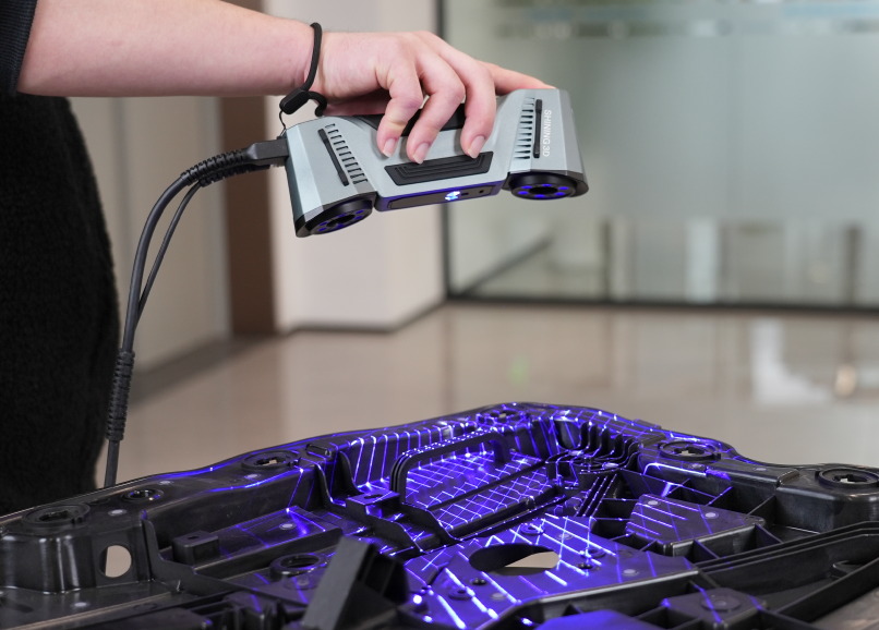 Handheld 3D scanner projecting blue laser lines across a large black part