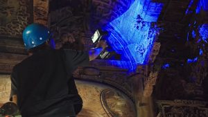 Handheld 3D scanner projecting blue light lines across an intricately carved timber panel in a heritage building