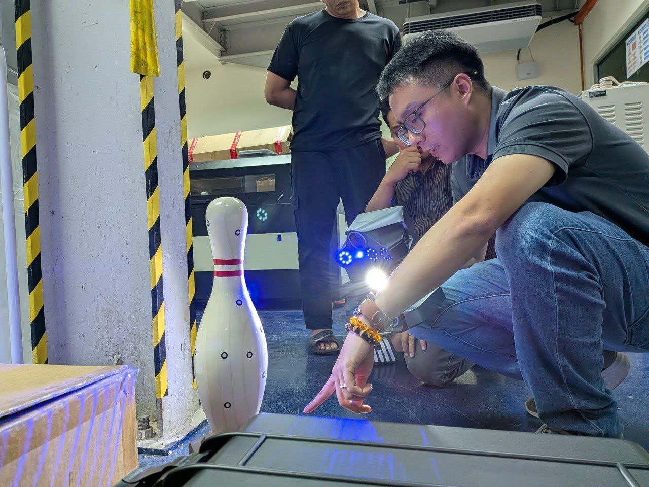 Engineer demonstrating a handheld 3D scanner on a sample part with a laptop on the floor in front of colleagues