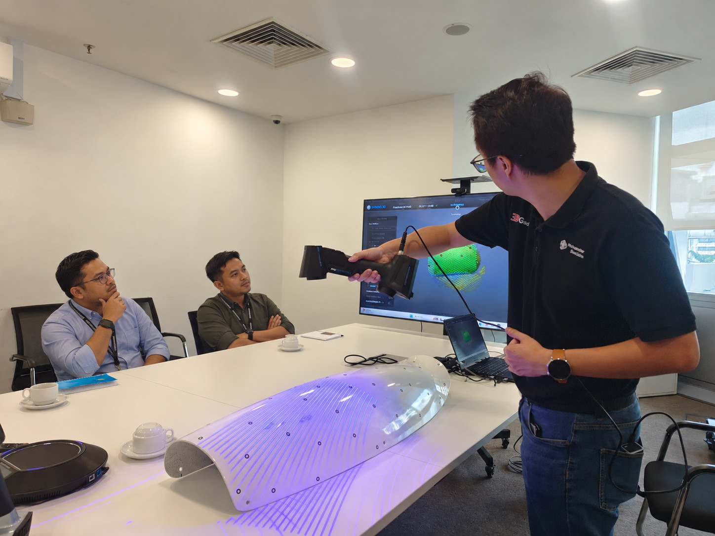 Handheld 3D scanner held over a curved panel during a live demo, scan building on the monitor behind