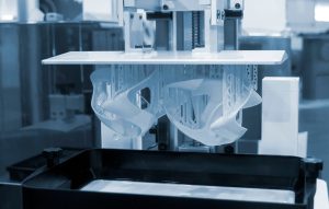 A desktop SLA resin 3D printer mid-build with the build platform lifted and the printed part hanging above the resin vat
