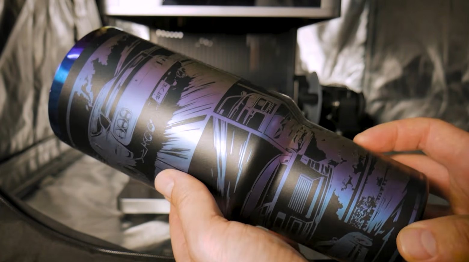 A black coated stainless steel tumbler engraved with a comic-book style design around its full circumference, held in the rotary attachment of a laser engraver