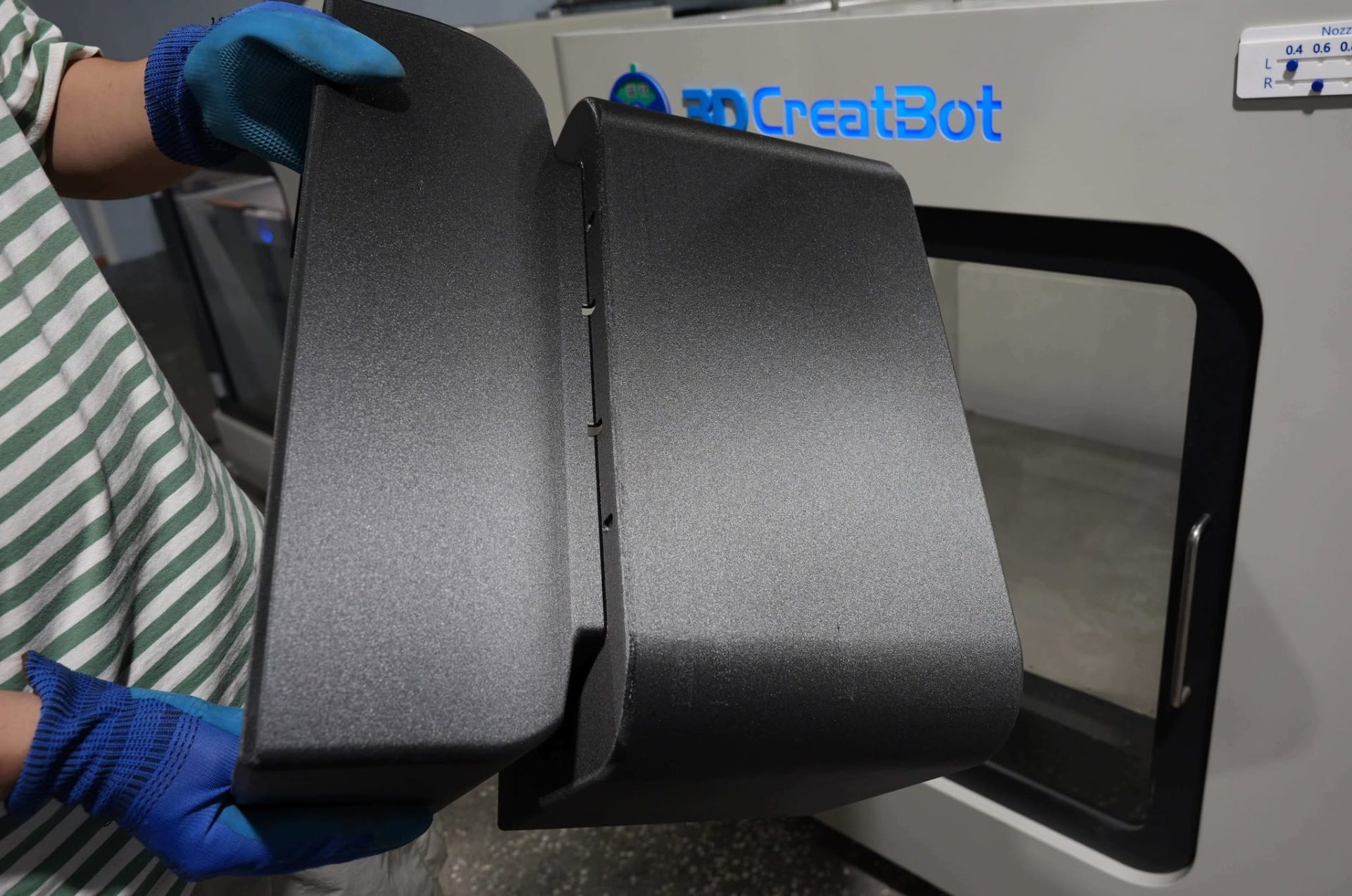 Gloved hands holding a large 3D printed black panel in front of a CreatBot printer