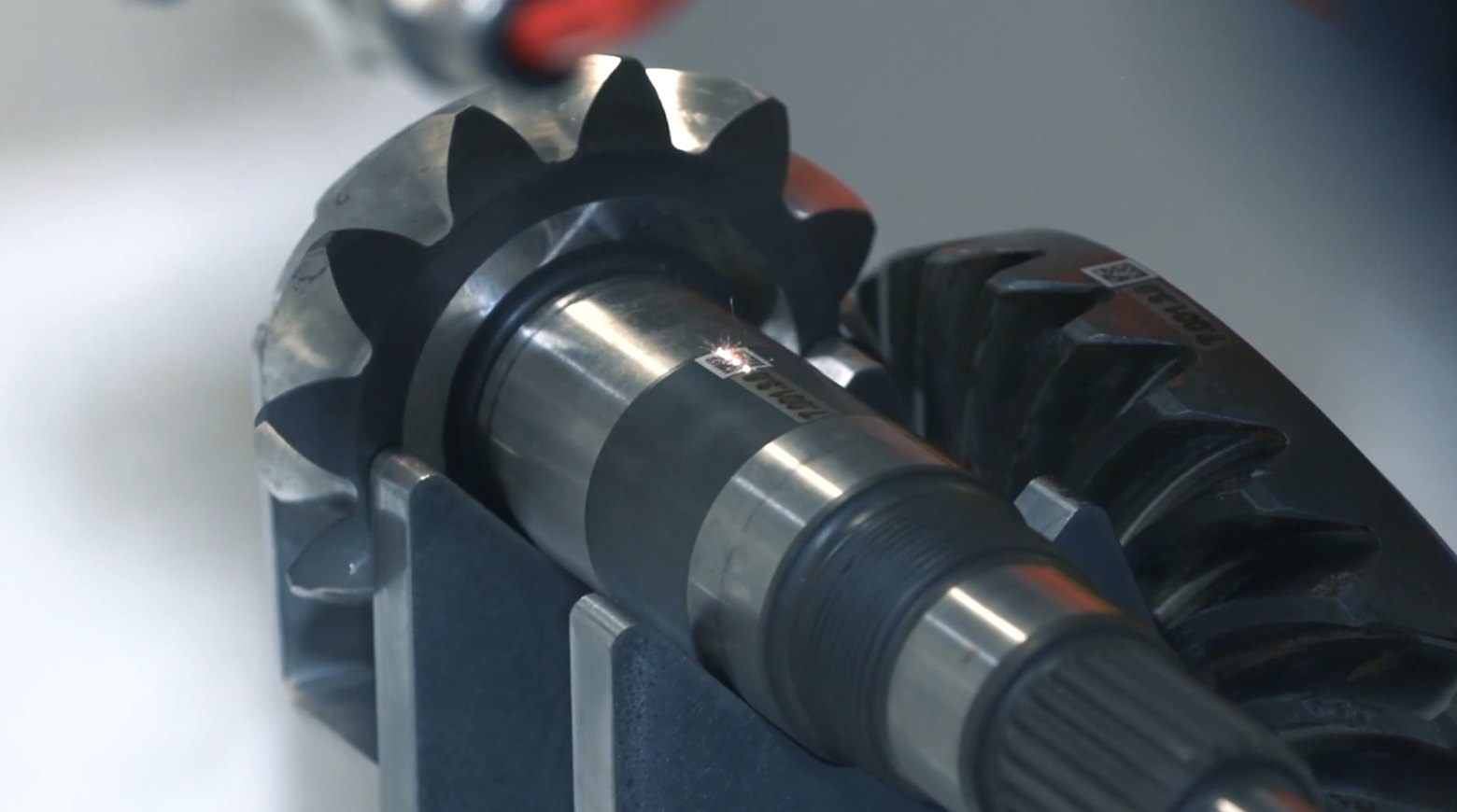 A laser marking a code onto the shaft of a bevel gear assembly held in a fixture, with the marking flash visible at the surface