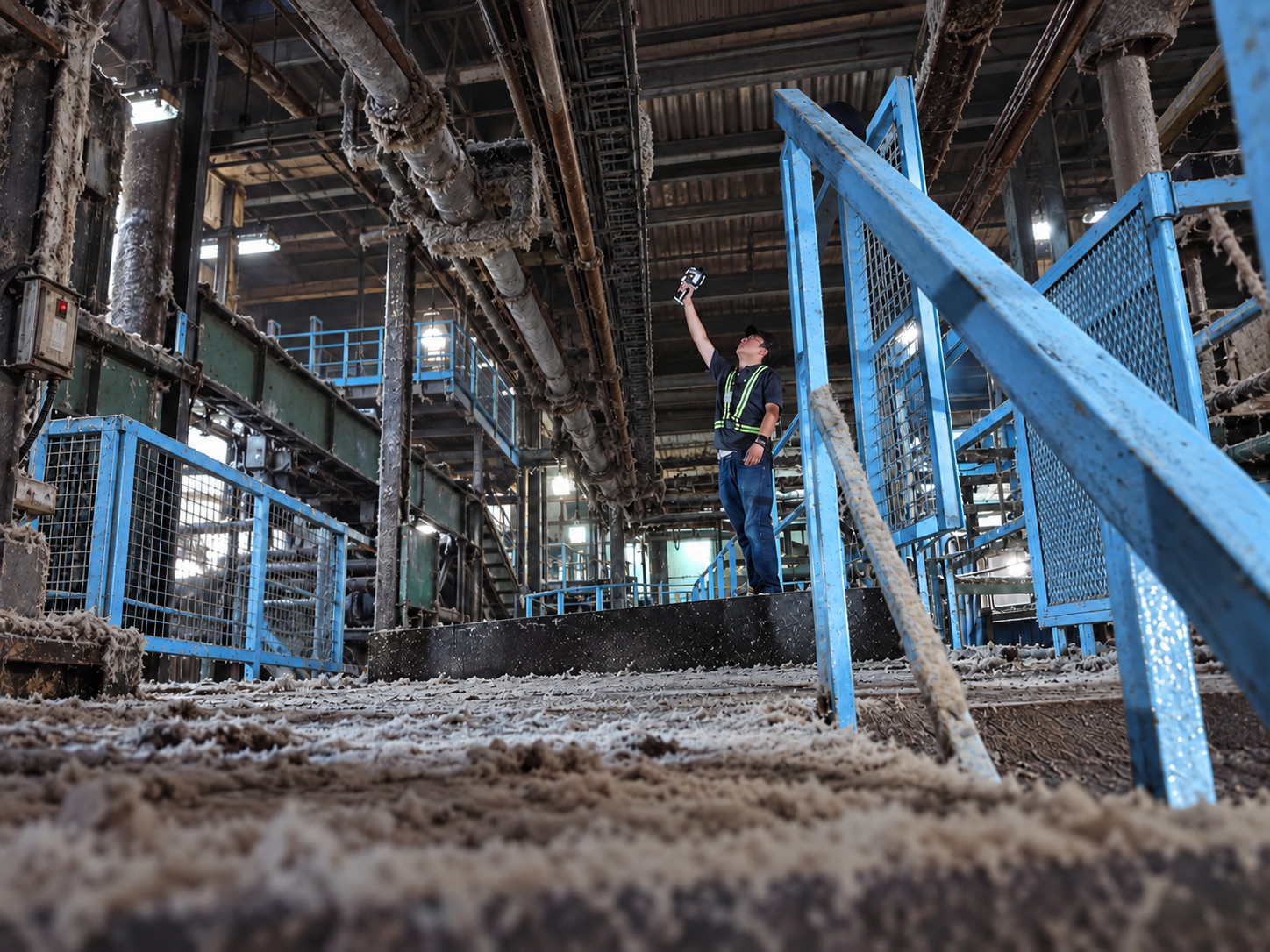 Worker in a hi-vis vest holding a handheld LiDAR scanner up towards pipework inside a plant