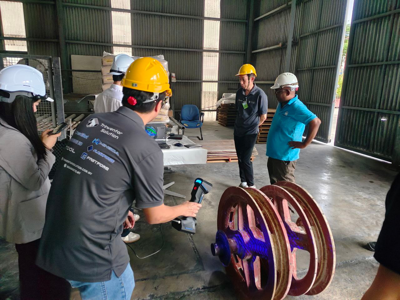 Two engineers using a handheld 3D scanner on large cast-iron wheels in a warehouse, with site staff watching