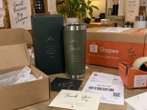 A personalised tumbler and gift box beside an open Shopee shipping carton, packing tape and a thank-you card