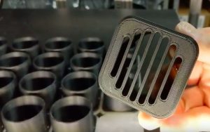 A finished PETG 3D printed part held up in front of a production batch of printed components