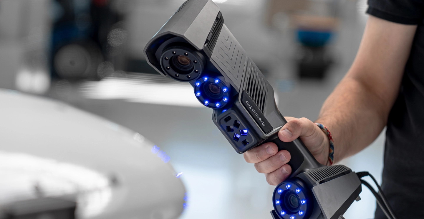 A handheld 3D scanner held in one hand, projecting blue light onto a car body panel