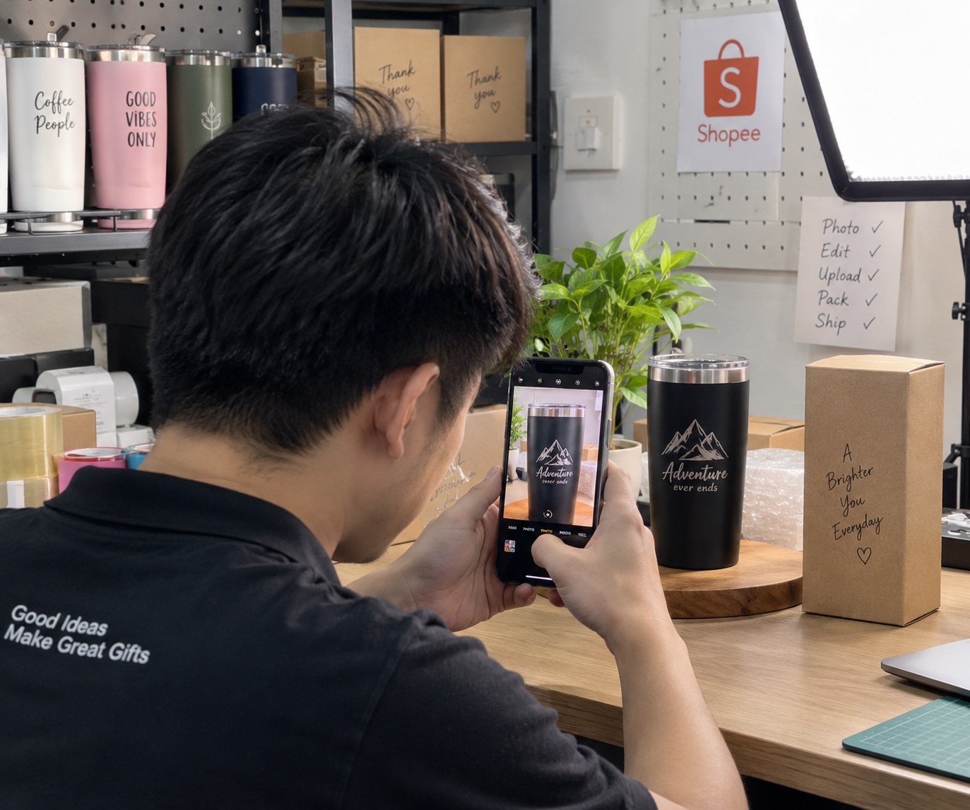 A seller photographing a personalised tumbler with a phone on a turntable, lit by a ring light at a packing bench