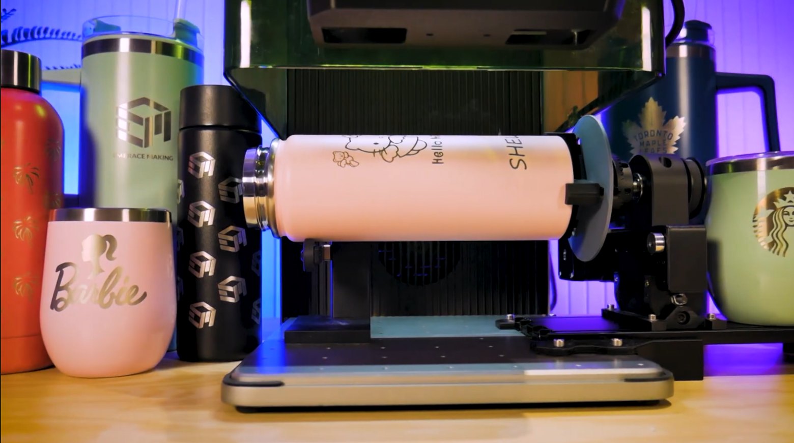 A tumbler held in a rotary attachment inside a laser engraver, surrounded by other engraved drinkware