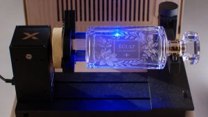 A perfume bottle held in a rotary attachment inside a UV laser, with a floral design and the name Eclat being engraved