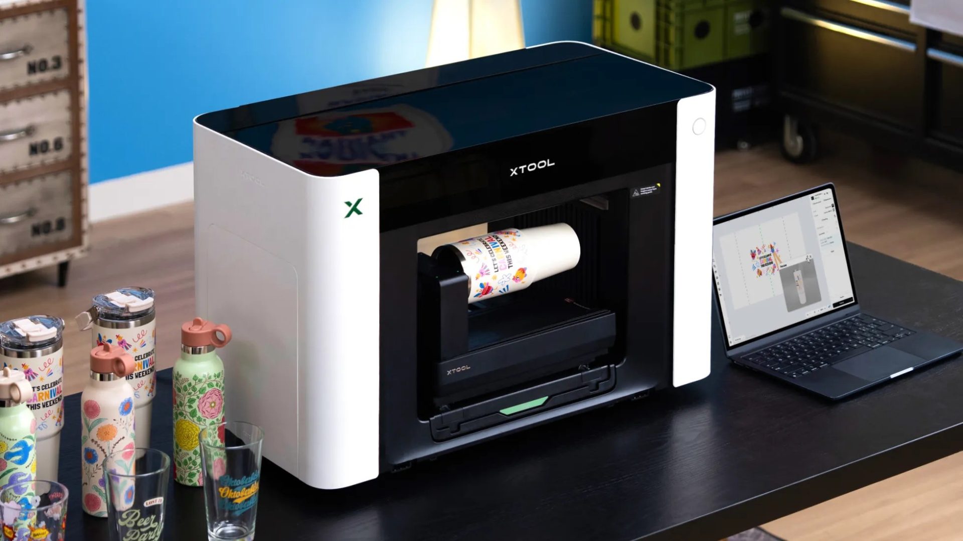The xTool O1 Omni Printer applying a full-colour design to a tumbler, with the artwork open on a laptop beside it